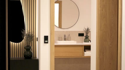 Ein Spiegel im Badezimmer mit Holzrahmen, daneben eine Vase mit Zweigen, dunkle Wandpaneele im Hintergrund.