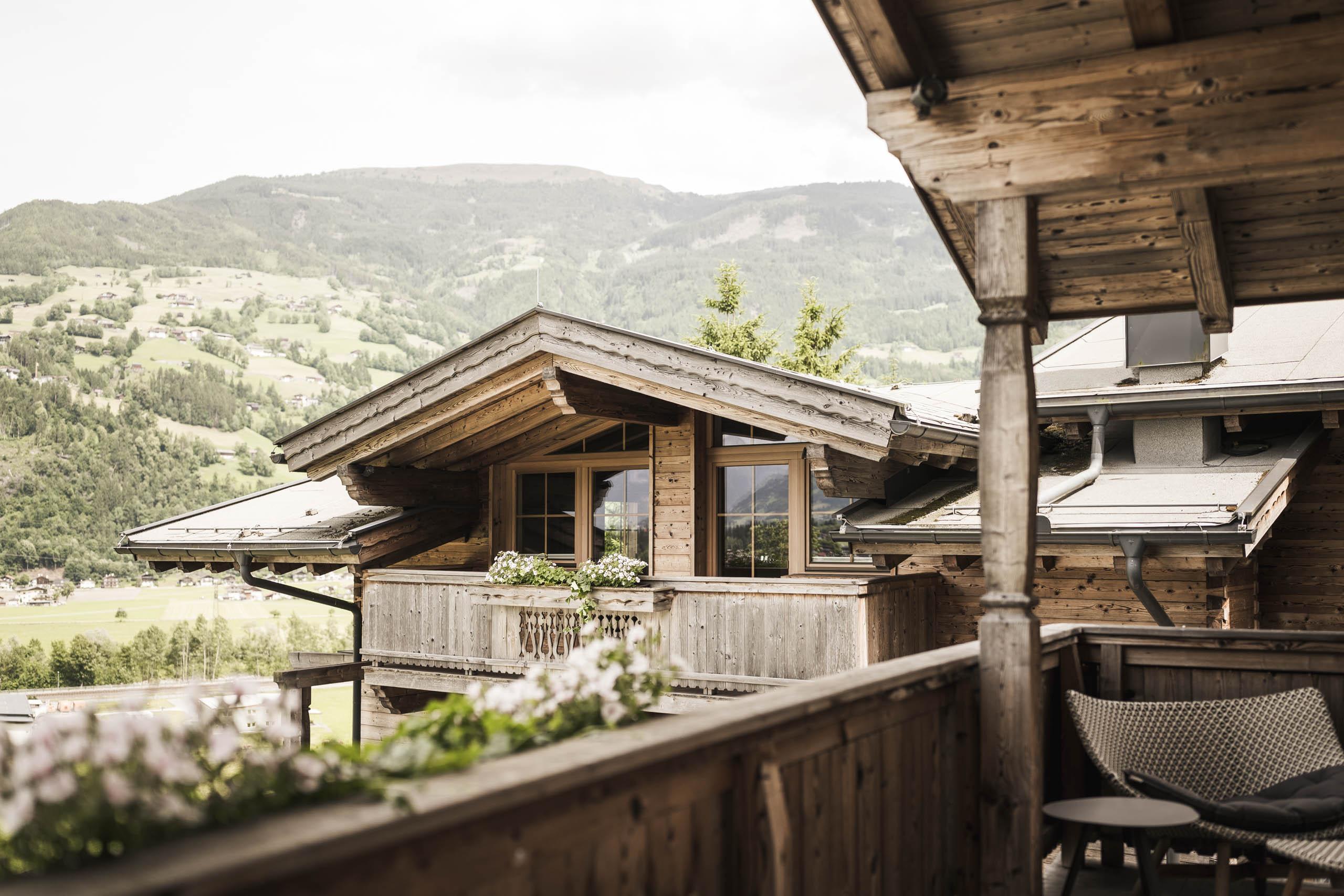 Balcony with wooden furniture and flowers, overlooking green hills and mountains in the background.