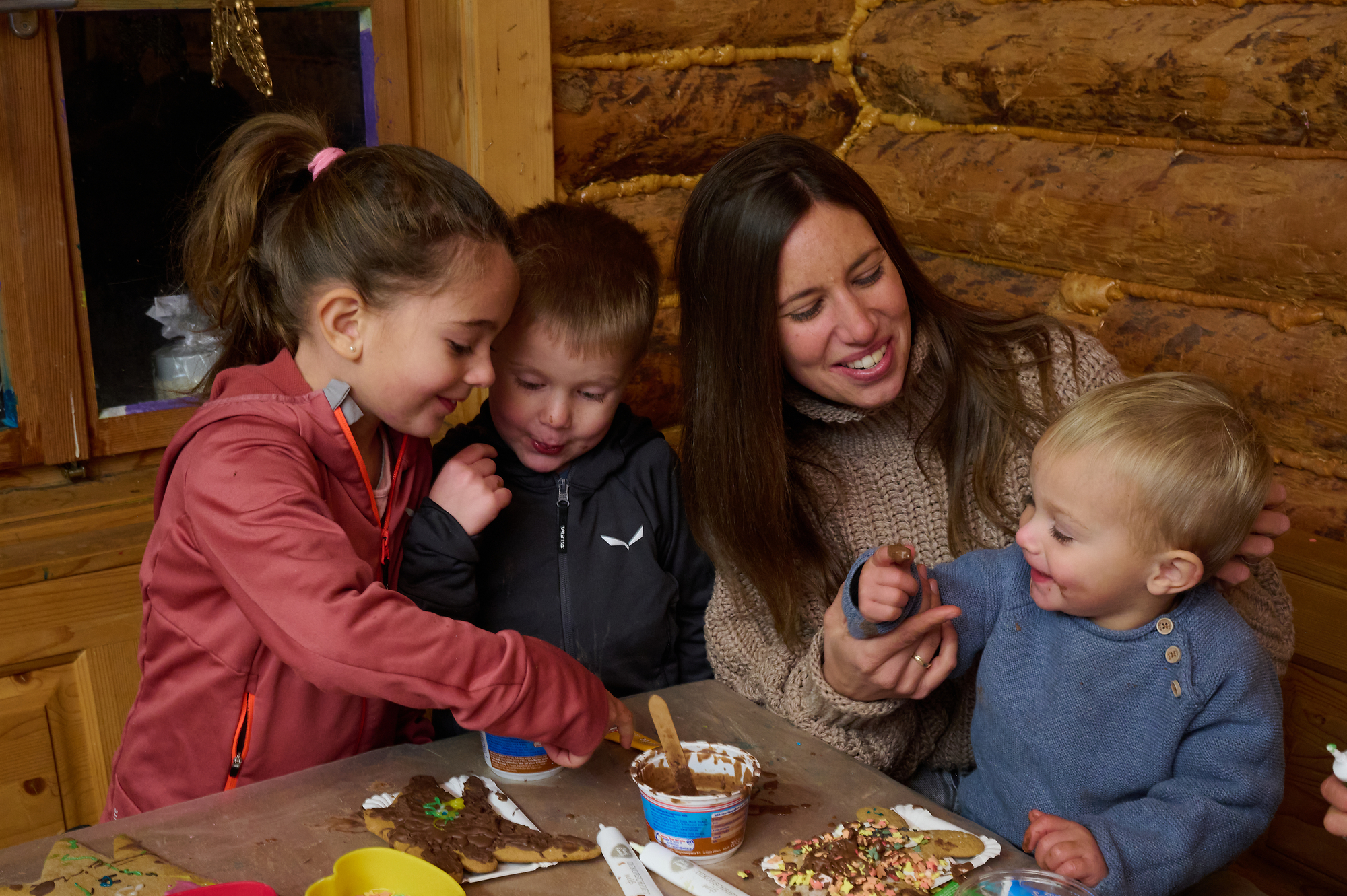 A woman sits at a table with three children and decorates gingerbread with chocolate.