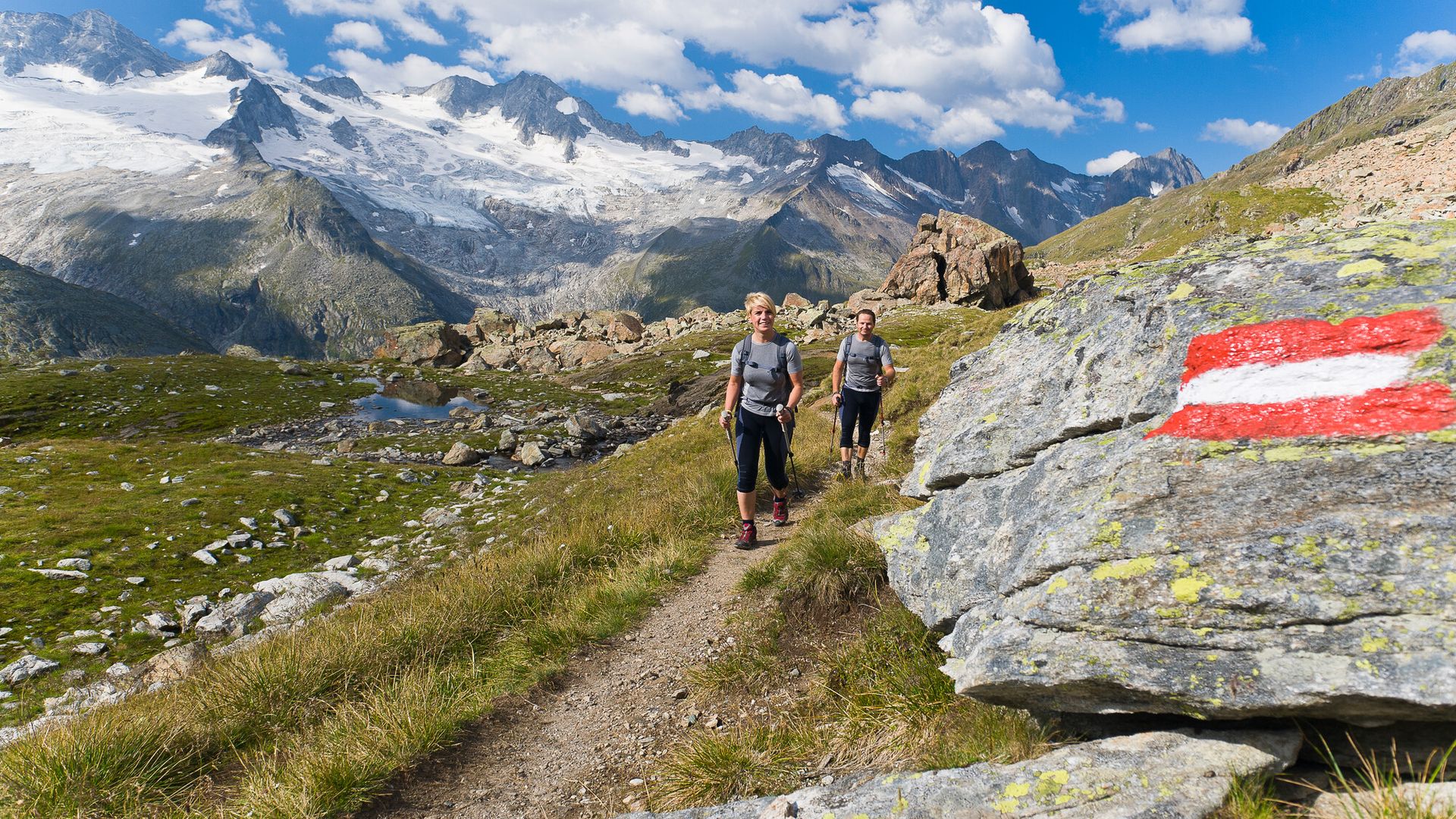Two hikers on the Berliner Höhenweg trail in the Zillertal valley. A marked rock shows the way, surrounded by green alpine meadows, rocks, and a clear mountain lake. In the background, mighty peaks rise up under a blue sky.