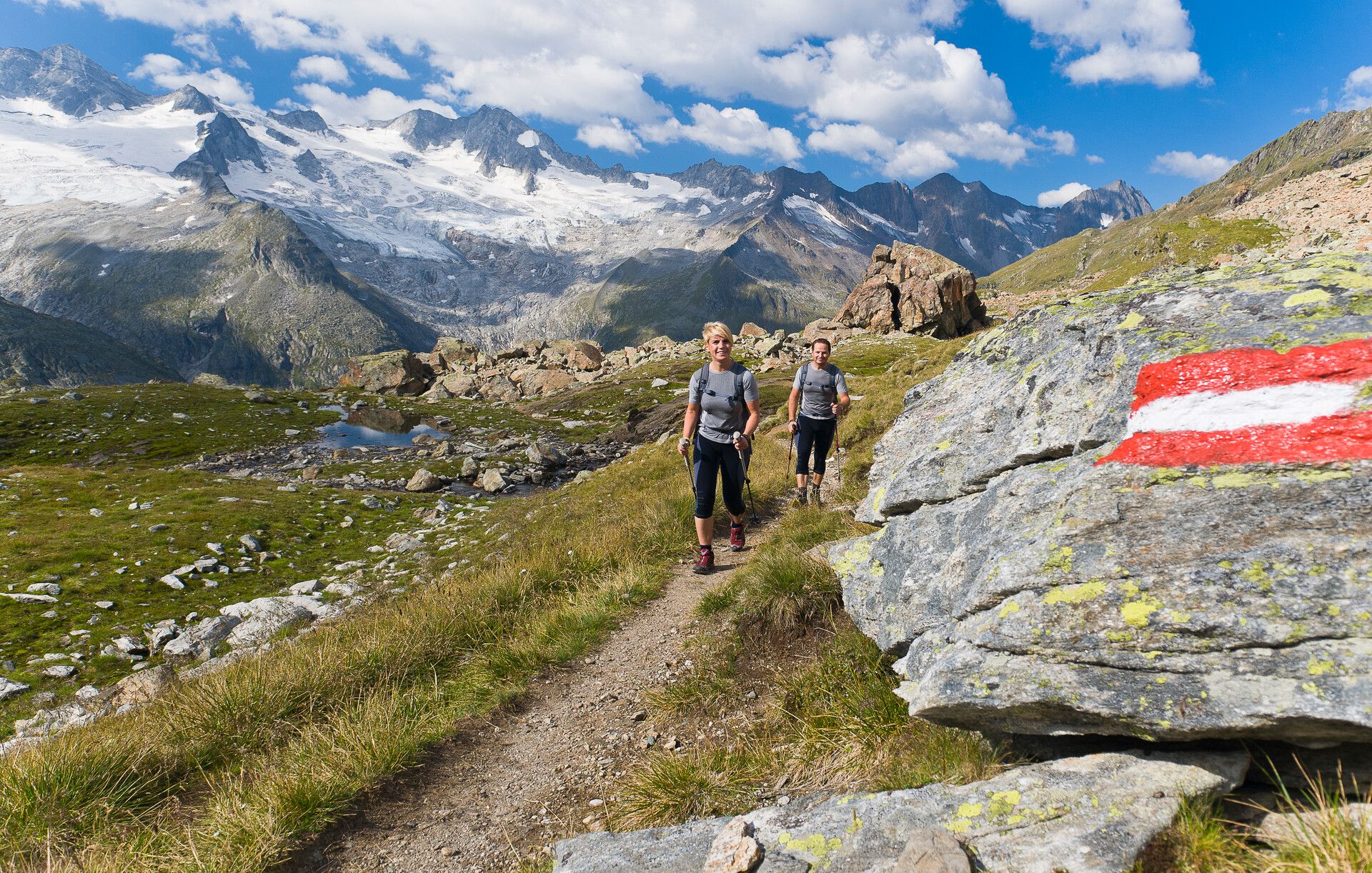 Two hikers on the Berliner Höhenweg trail in the Zillertal valley. A marked rock shows the way, surrounded by green alpine meadows, rocks, and a clear mountain lake. In the background, mighty peaks rise up under a blue sky.