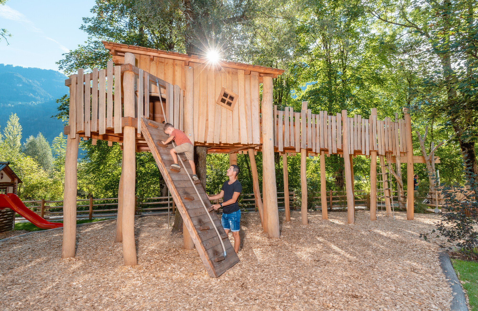 A child climbs a slanted wooden ramp to a treehouse at the Auenland Sidan playground in Schwendau. The playhouse is surrounded by trees, with sunlight in the background and mountains in the distance.
