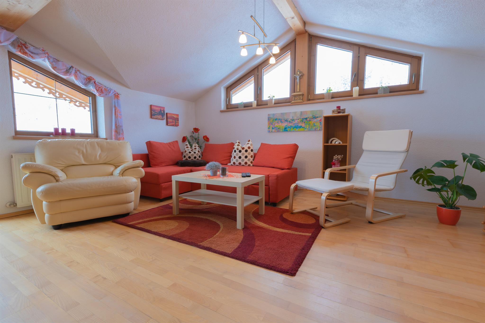 Heller Wohnzimmerbereich mit Sofa, Sessel, Couchtisch, Wandbilder, Pflanzen und großen Fenstern unter Dachschräge.