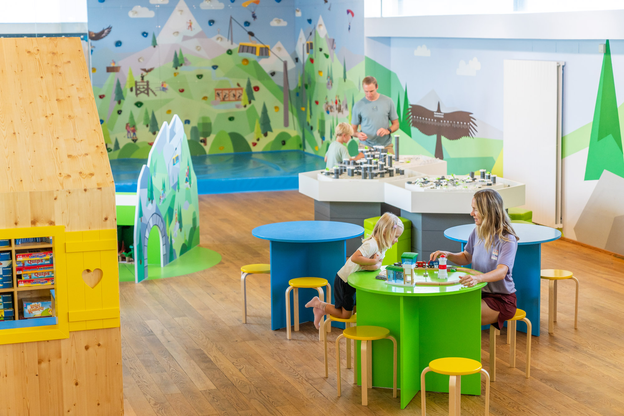 Kinder und Erwachsene spielen in einem bunten, kindgerechten Raum mit grünen Tischen und Wandmalereien.