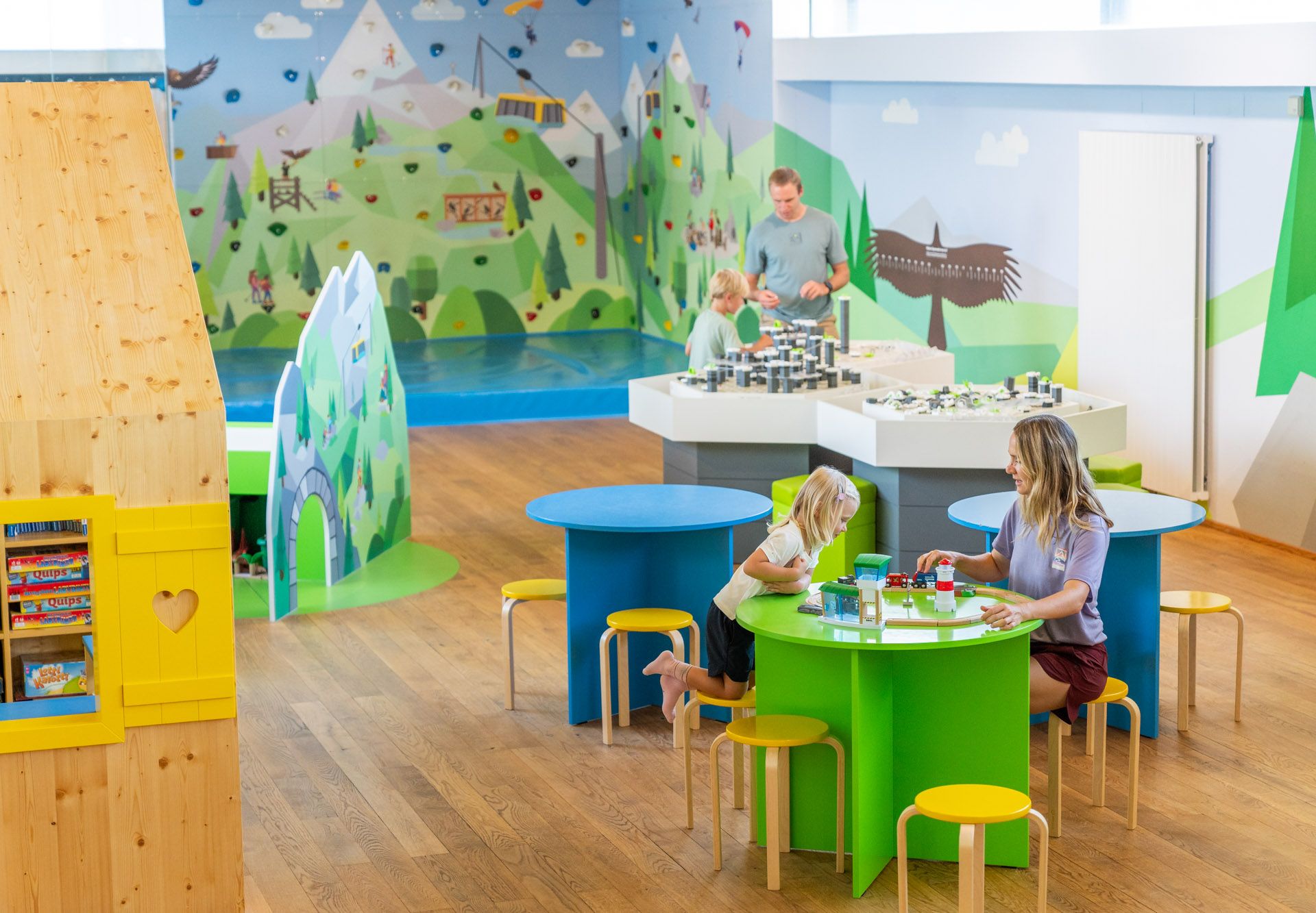 Kinder und Erwachsene spielen in einem bunten, kindgerechten Raum mit grünen Tischen und Wandmalereien.