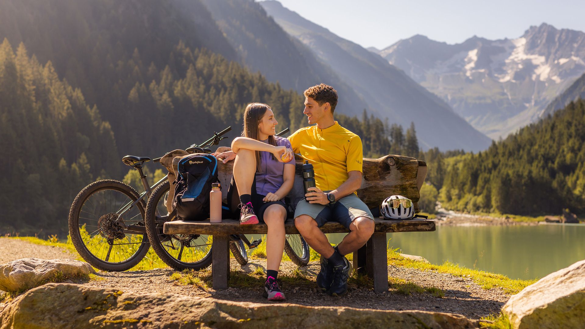 Two cyclists take a break on a bench in Stilluptal, Zillertal. In the background: the Stillup reservoir, green forests and imposing mountains in the sunshine.