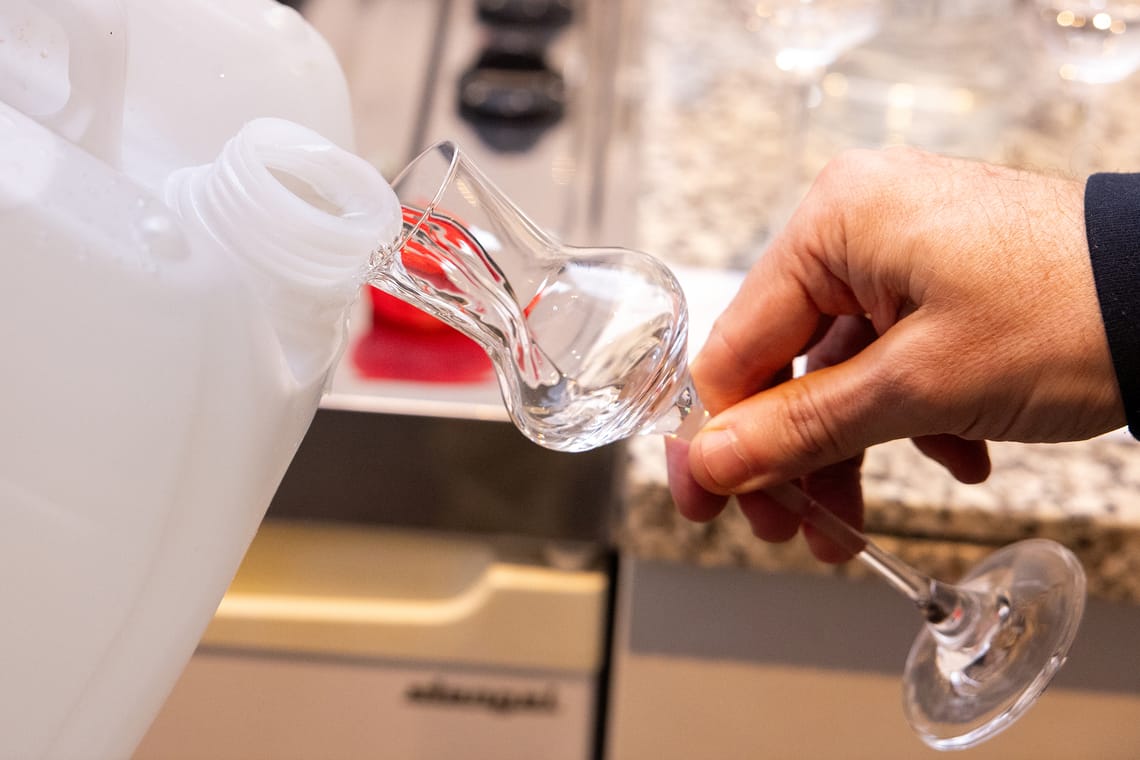 HIPPSTAR Abfüllung – Brennmeister füllt Edelbrand vom Kanister ins Glas | Mayrhofen-Hippach Eine Hand füllt den HippStar-Edelbrand aus einem großen weißen Kunststoffkanister in ein Schnapsglas mit langem Stiel ab, im Hintergrund unscharf eine Küchenzeile.