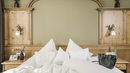 Bett mit weißen Kissen, Nachttisch mit Telefon, Vase und Tasche, Holzkopfteil mit Lampen an der Wand.