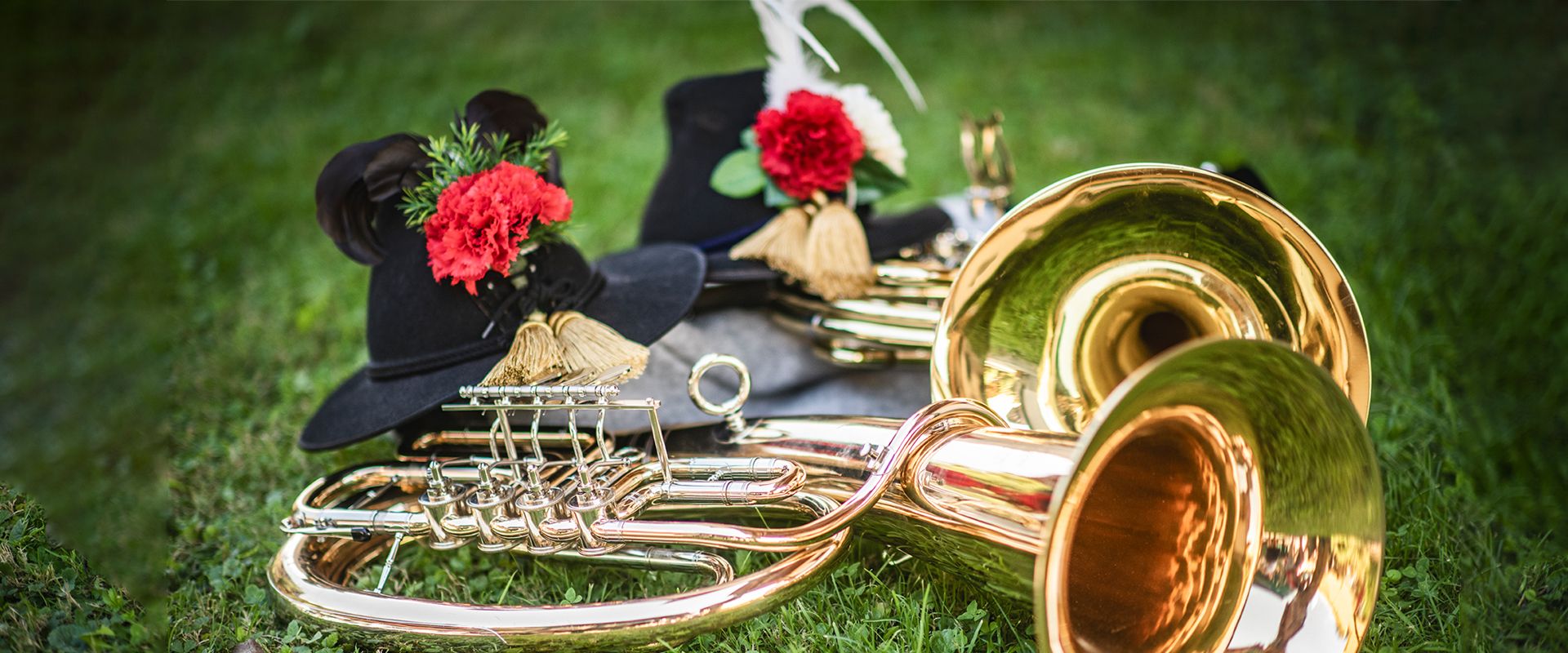 Blasmusikinstrumente und traditionelle Hüte mit Federn und roten Blumen liegen auf einer grünen Wiese. Das Bild symbolisiert die alpenländische Musikkultur in Mayrhofen-Hippach im Zillertal.