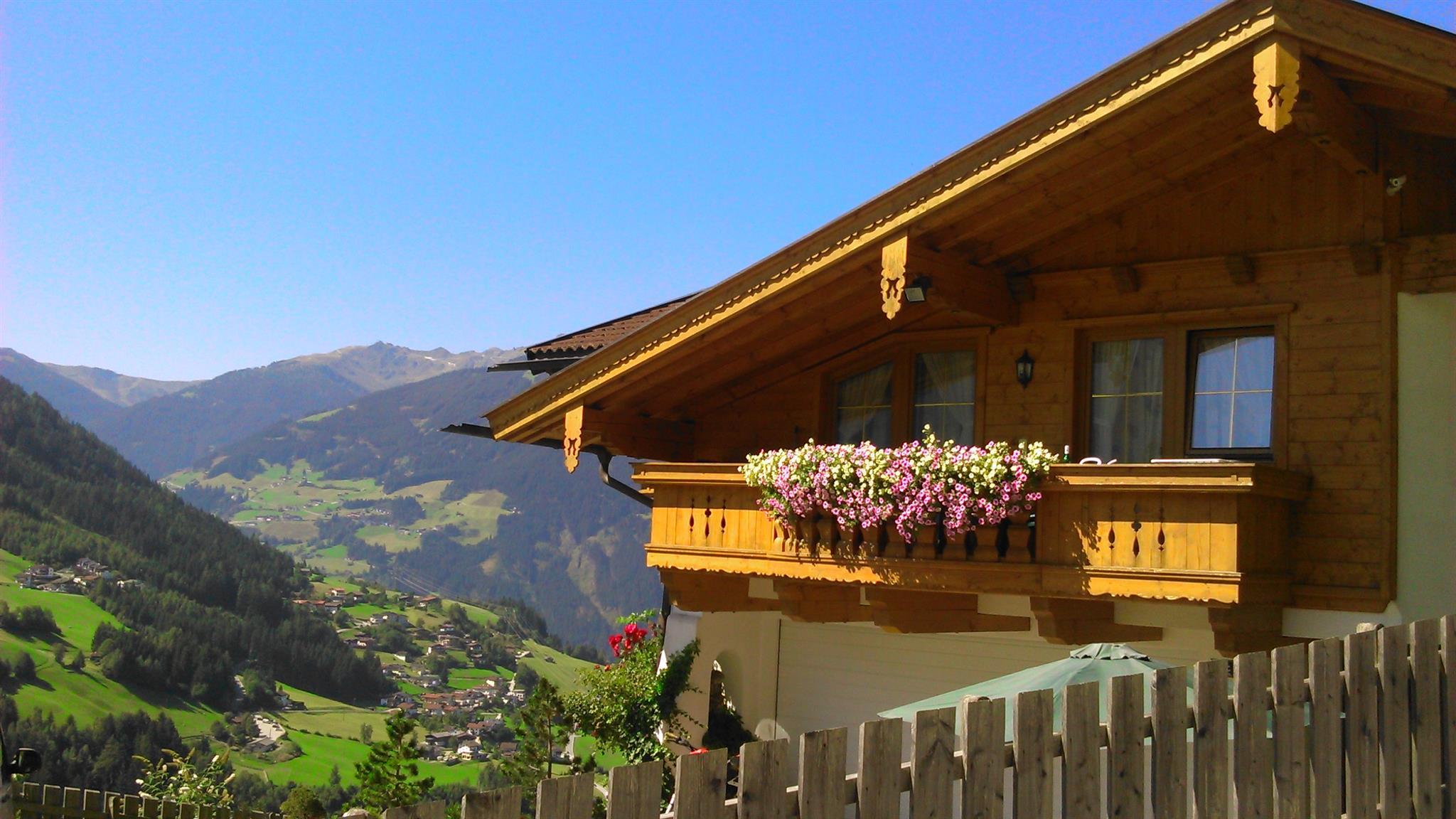 Häuser im alpinen Stil mit Holzfassaden und Balkon, vor grünen Hügeln und Bergen bei klarem Himmel.