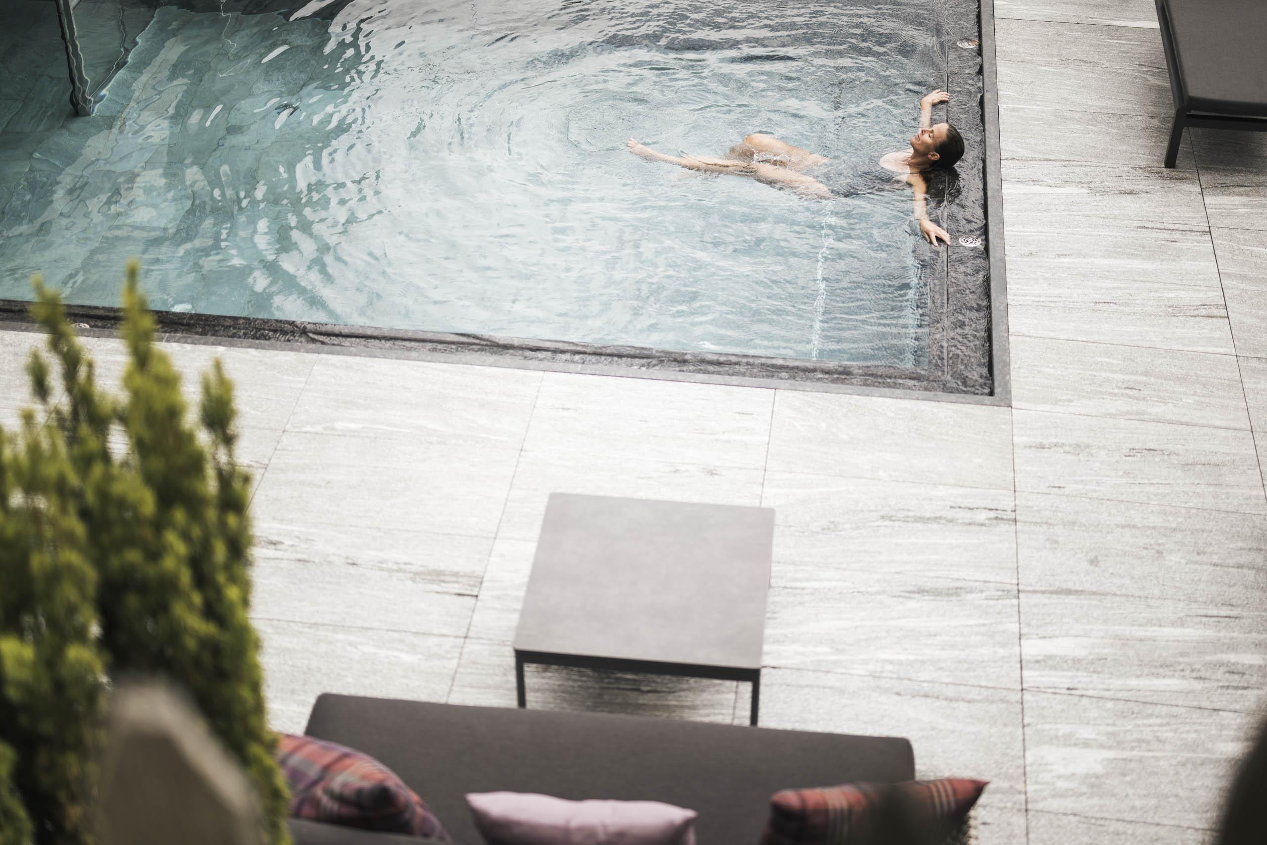 Woman relaxing in a pool, with dark hair and closed eyes, near the pool edge and furniture.