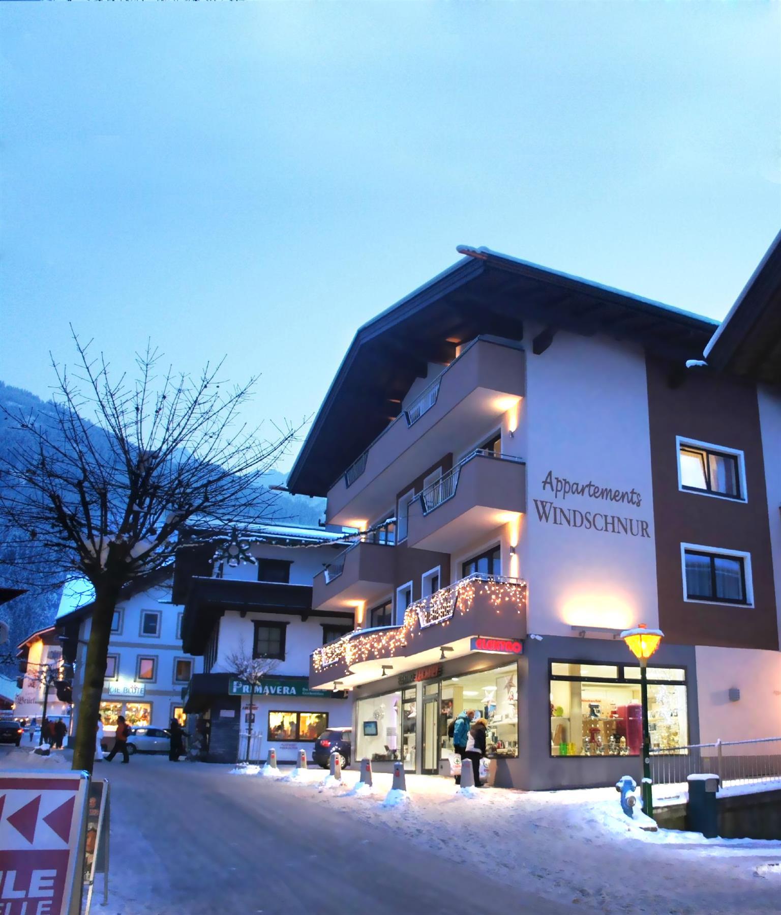 Multi-story building with Christmas lights, shops on the ground floor, snowy ground, evening sky, people walking.