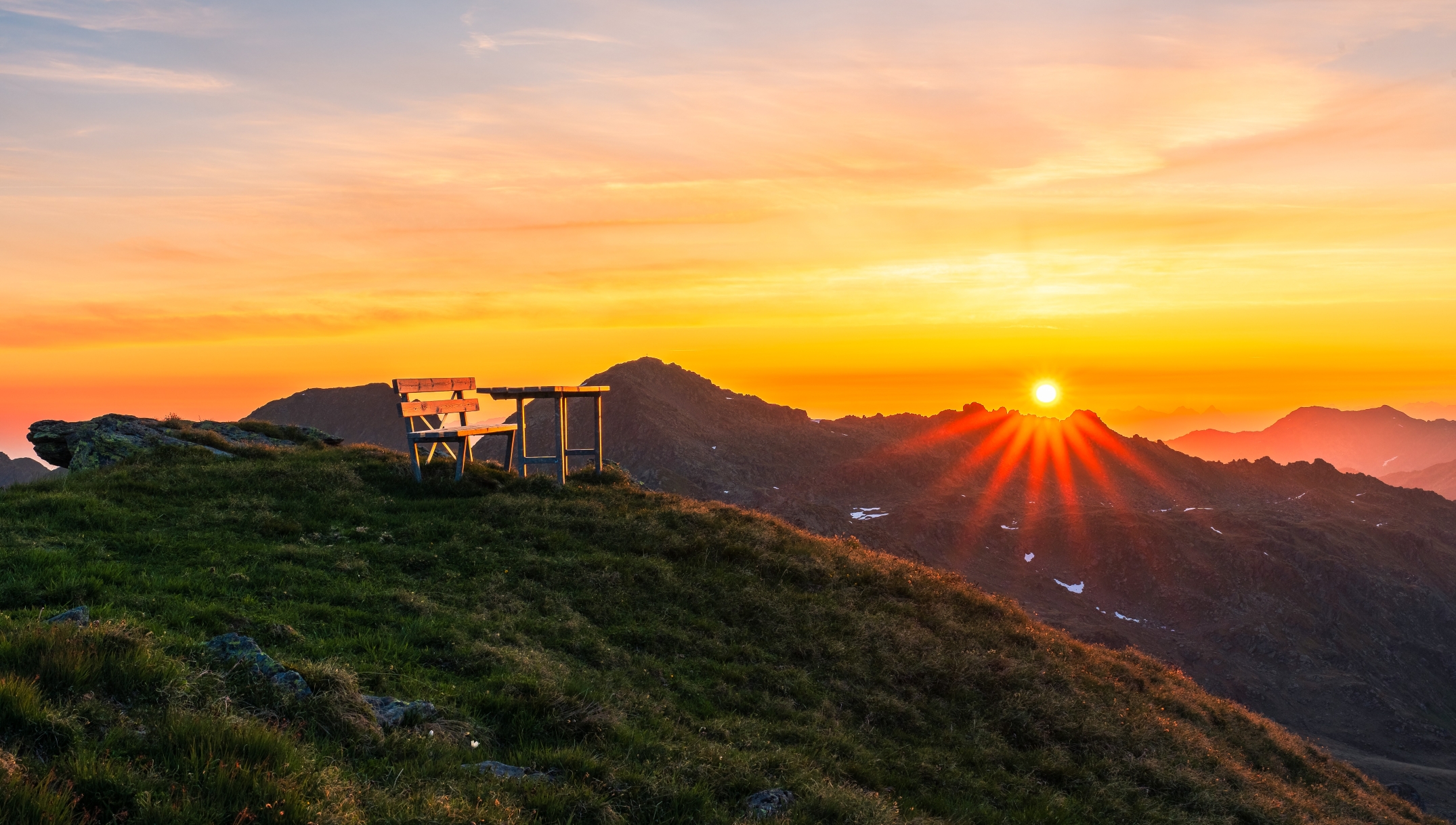 Vorne der Gipfel des Rifflerkogl mit einem Tisch und einer Bank zum Sitzen im Hintergrund der Sonnenaufgang 