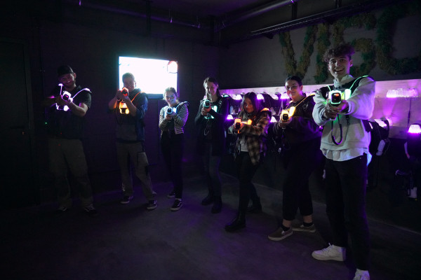 The image shows a group of seven people playing laser tag. Everyone is wearing bright vests and holding their laser tag guns towards the camera. The room is dark, but the colorful lights of the equipment shine brightly.