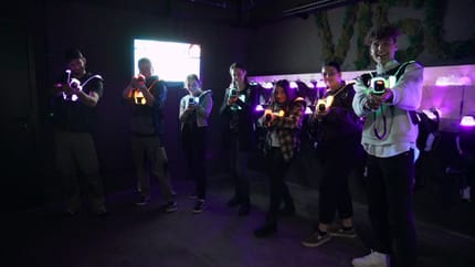 The image shows a group of seven people playing laser tag. Everyone is wearing bright vests and holding their laser tag guns towards the camera. The room is dark, but the colorful lights of the equipment shine brightly.