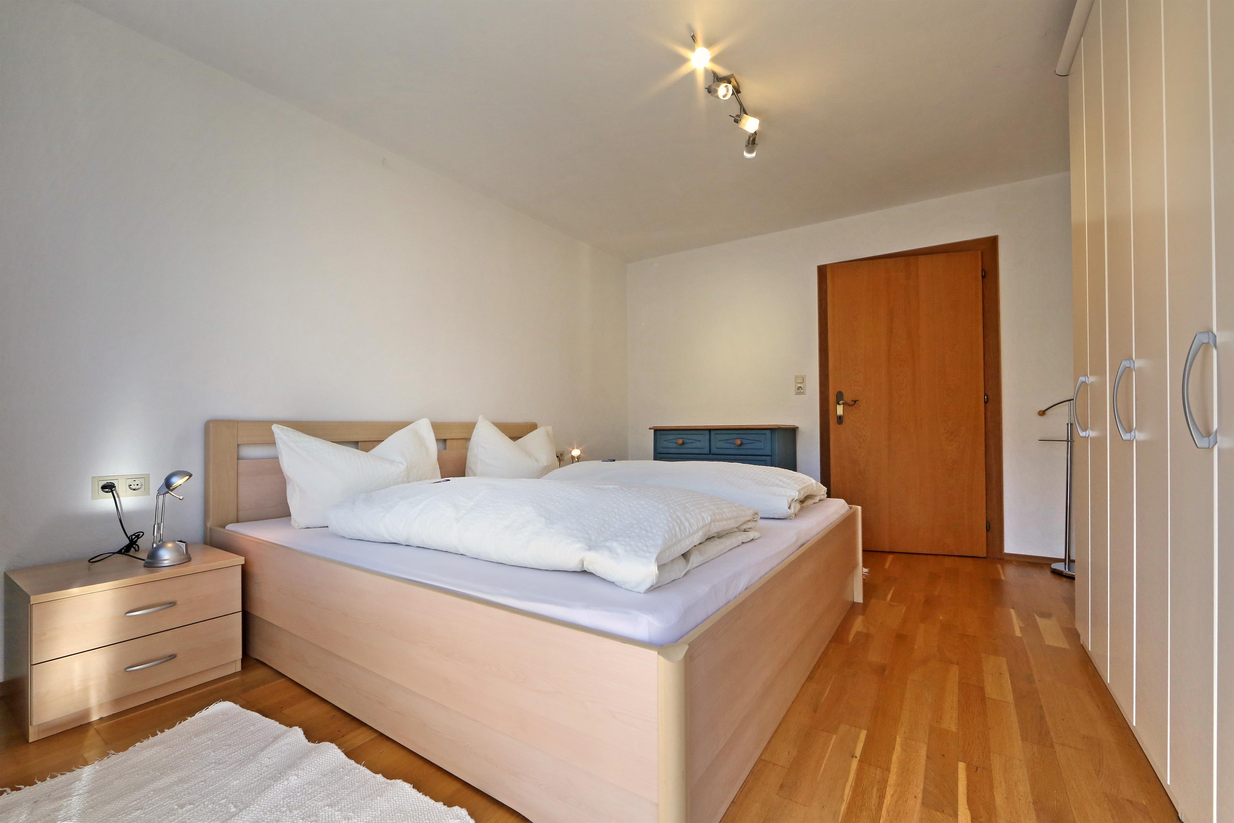 Schlafzimmer mit Doppelbett, Nachttisch, Leselampen, Holzfußboden, Tür und Kleiderschrank sichtbar.