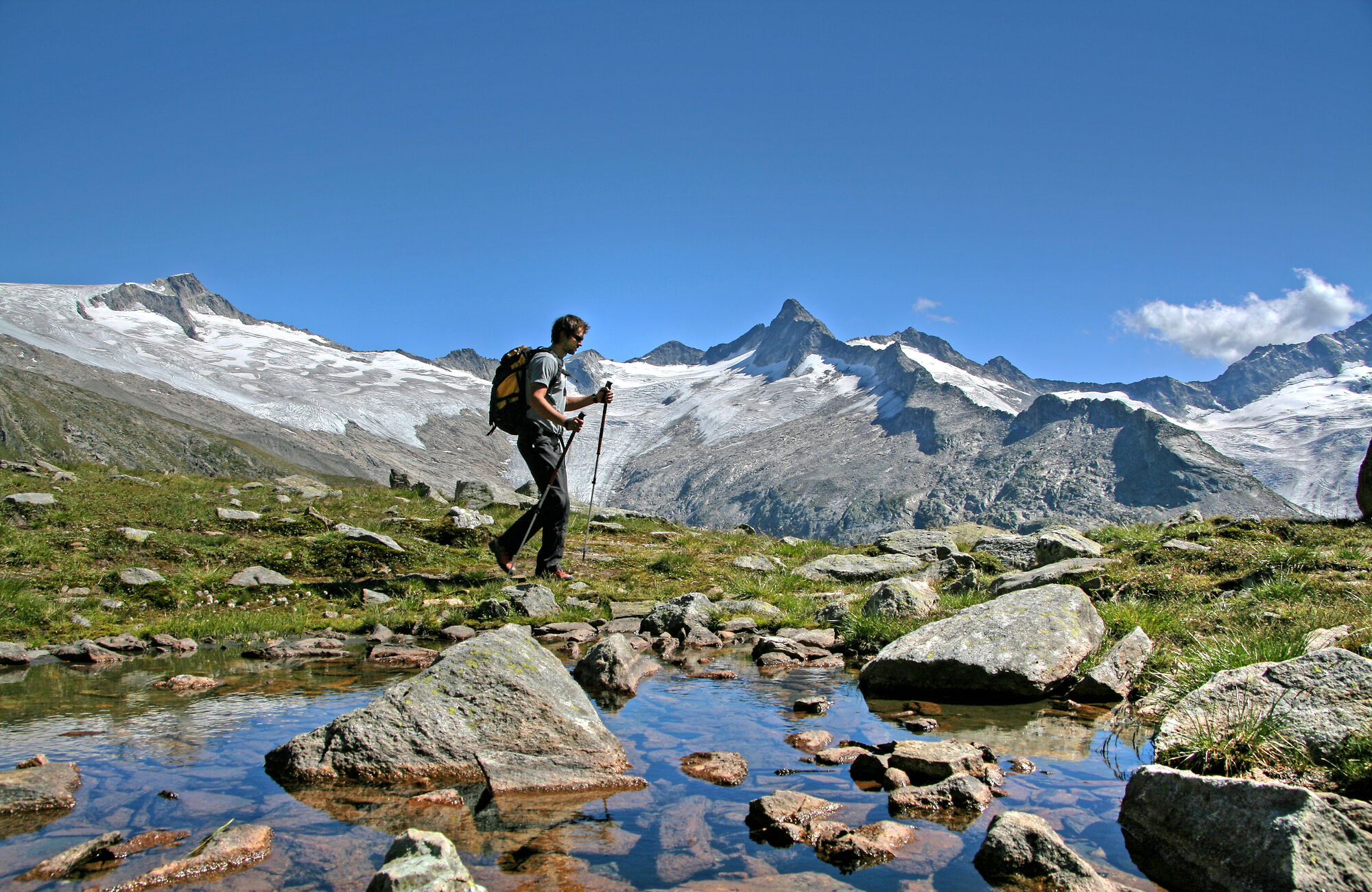 A hiker with a backpack crosses a stream on stones in an alpine meadow in the Zillertal, on the Berliner Höhenweg near Mayrhofen-Hippach, with snow-covered peaks under a blue sky.