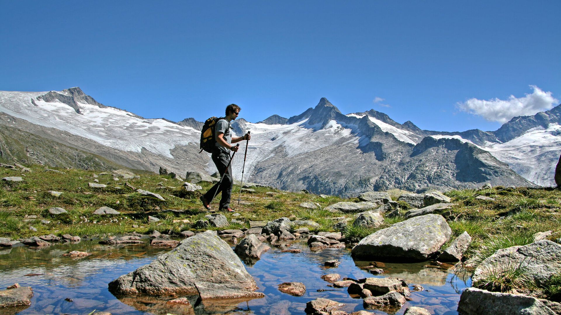 A hiker with a backpack crosses a stream on stones in an alpine meadow in the Zillertal, on the Berliner Höhenweg near Mayrhofen-Hippach, with snow-covered peaks under a blue sky.