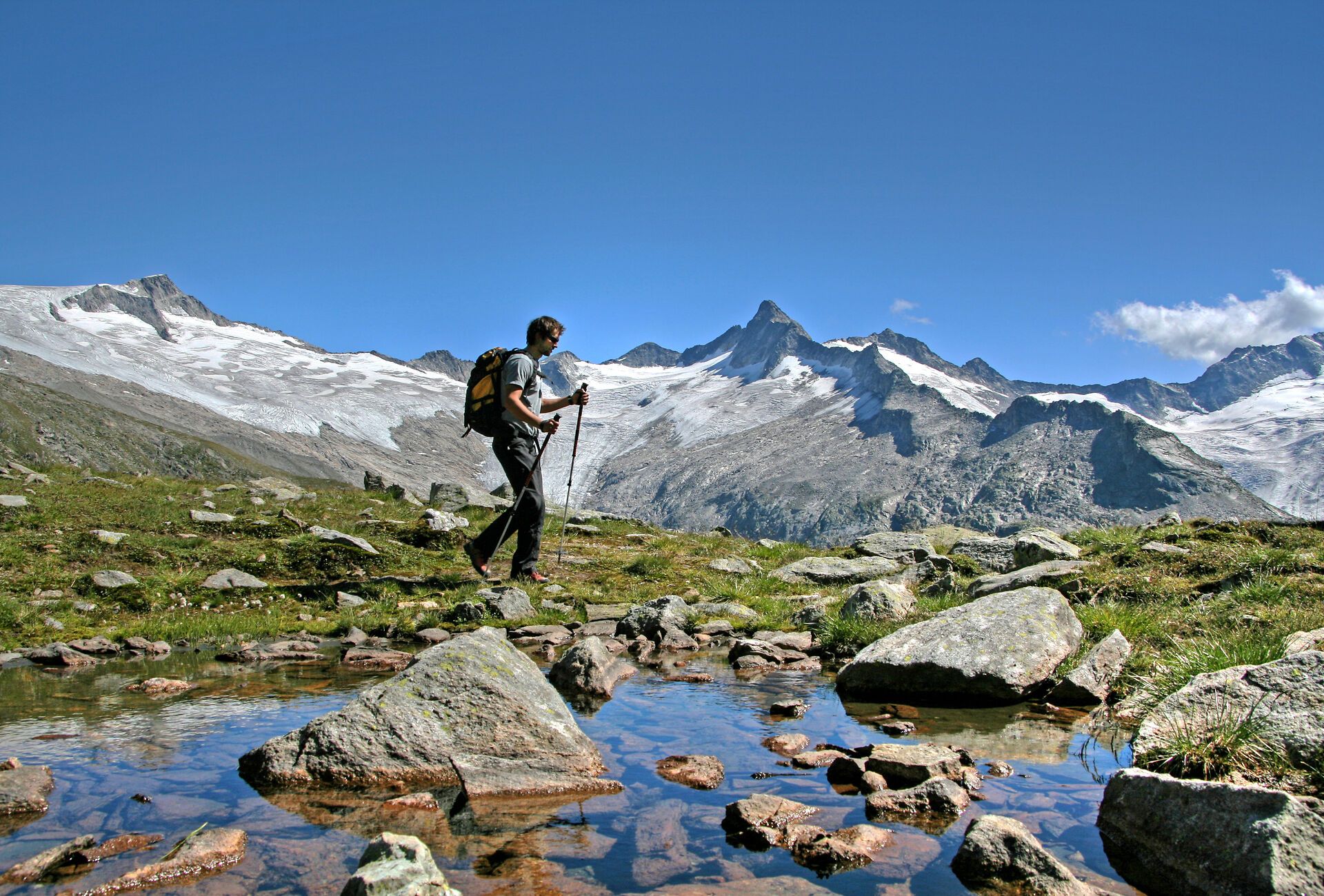 A hiker with a backpack crosses a stream on stones in an alpine meadow in the Zillertal, on the Berliner Höhenweg near Mayrhofen-Hippach, with snow-covered peaks under a blue sky.