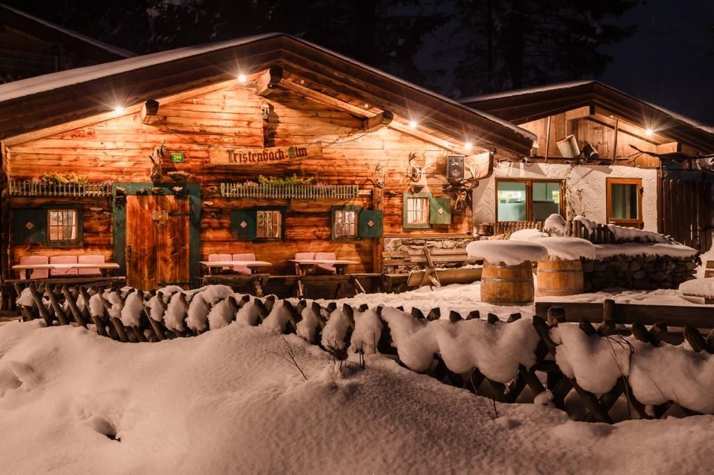 Eine urige, beleuchtete Almhütte mit einer Terrasse, die mit Schnee bedeckt ist