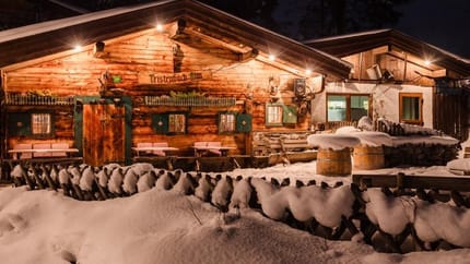 Eine urige, beleuchtete Almhütte mit einer Terrasse, die mit Schnee bedeckt ist