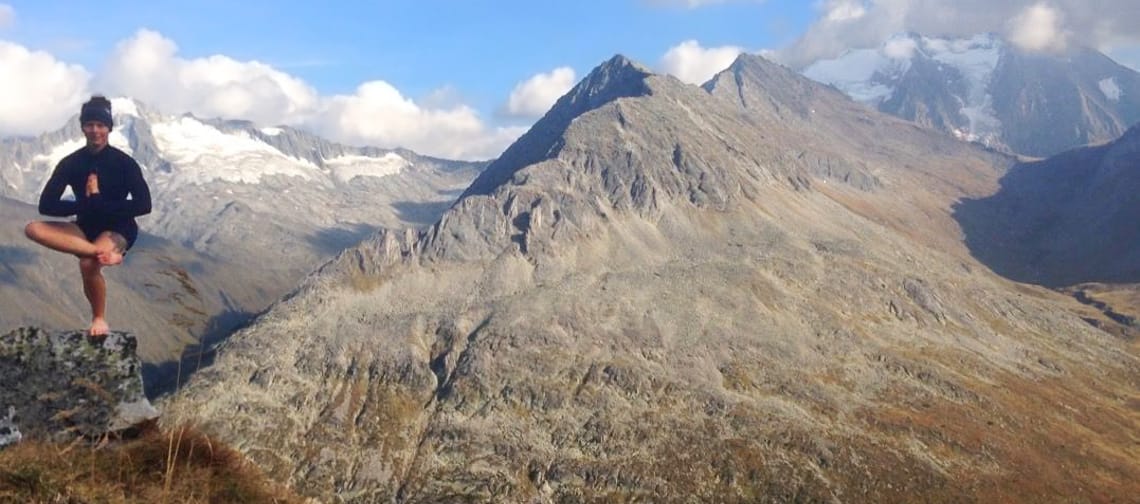 Eine Person macht eine Yogaposition auf einem Stein, im Hintergrund sieht man Berge