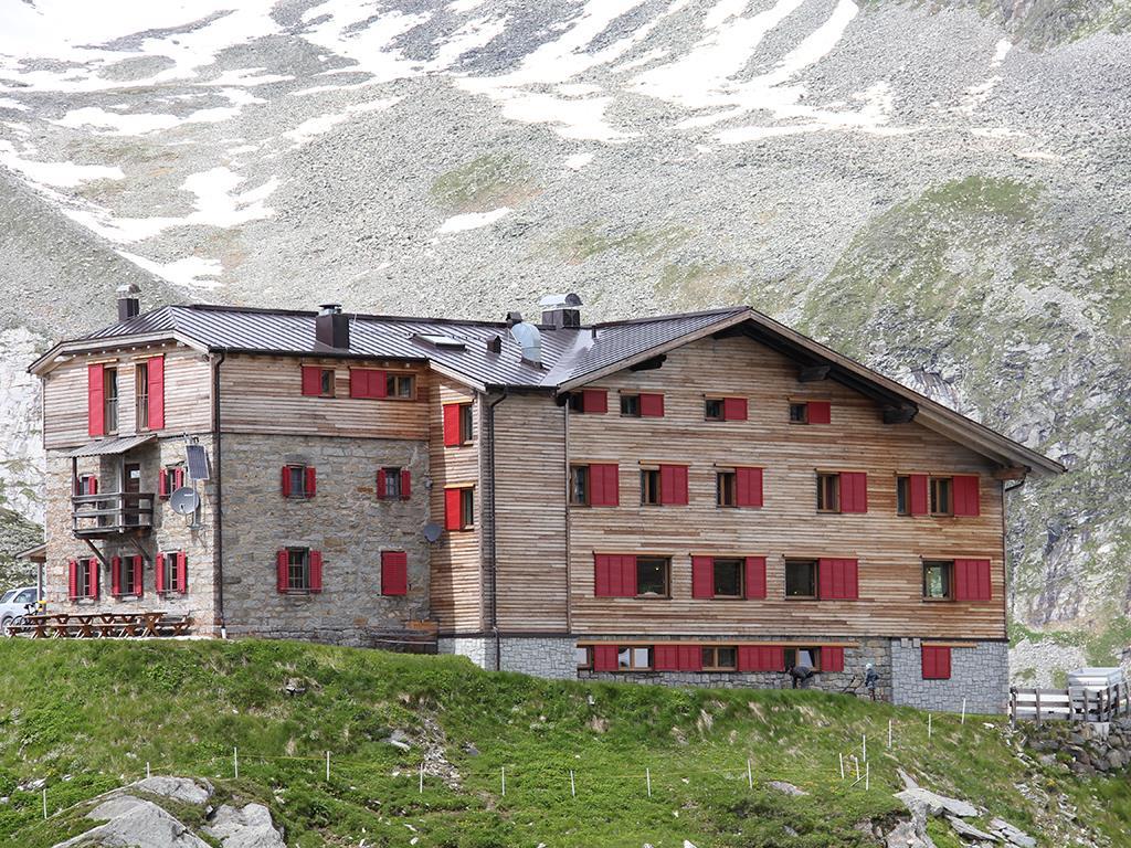 A multi-story mountain house with wooden exterior, red shutters, and green grass in front of a snowy mountain.