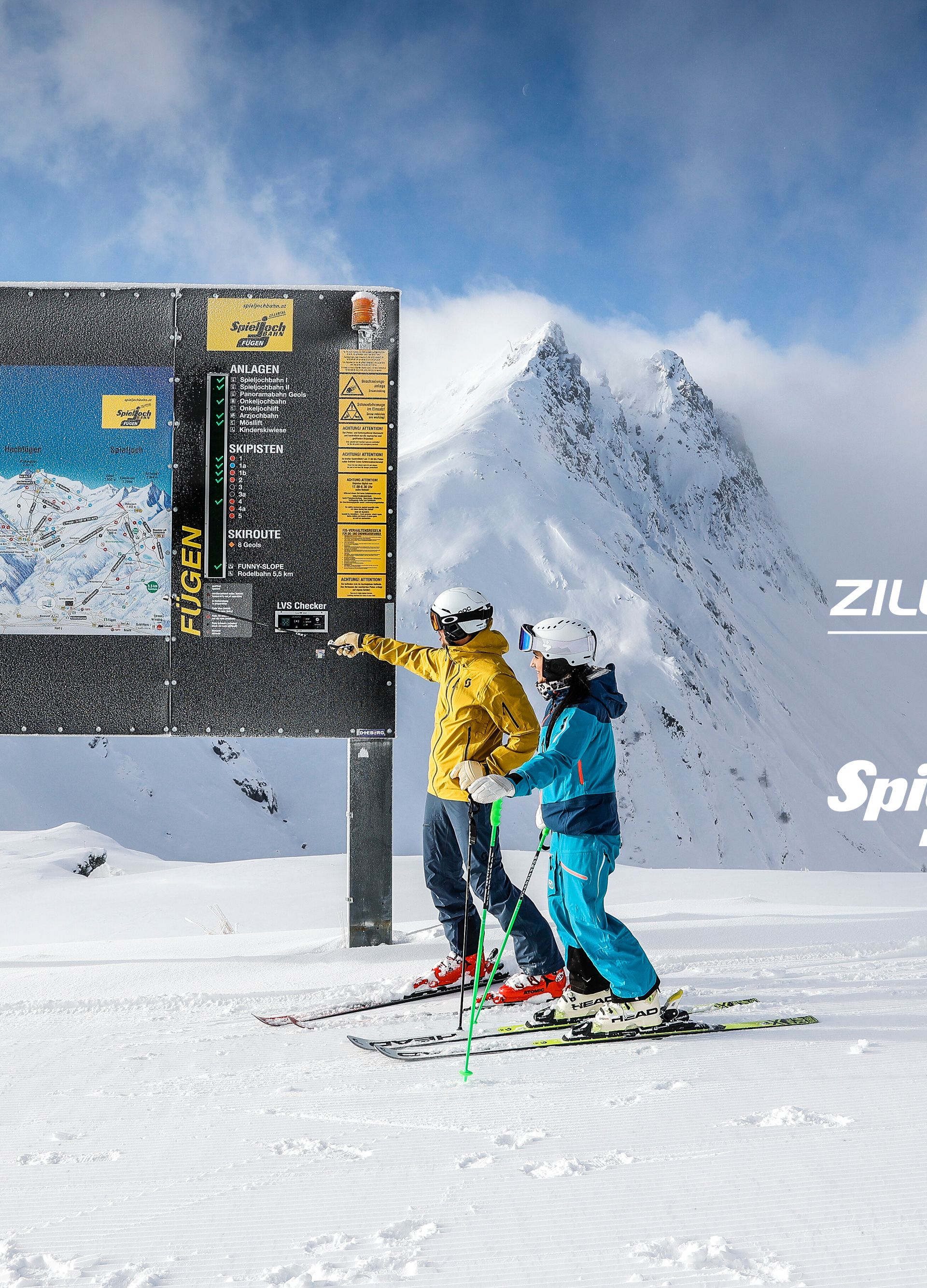Two skiers in front of a large trail map sign in snowy mountain scenery.