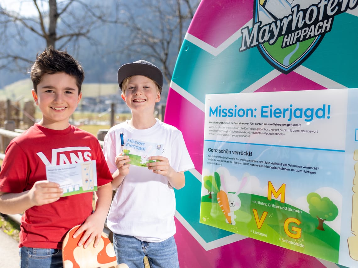 Mission Egg Hunt Mayrhofen-Hippach – Success at the Easter Egg Station | Tyrol Two children stand next to a large Easter egg from the “Mission Egg Hunt” in Mayrhofen-Hippach, showing their found clues; colourful riddle station in the background.