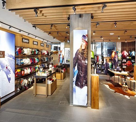 Innenansicht eines Sportgeschäfts mit Wandregalen voller Taschen und Kleidung, Holzdecke und digitaler Werbetafel.