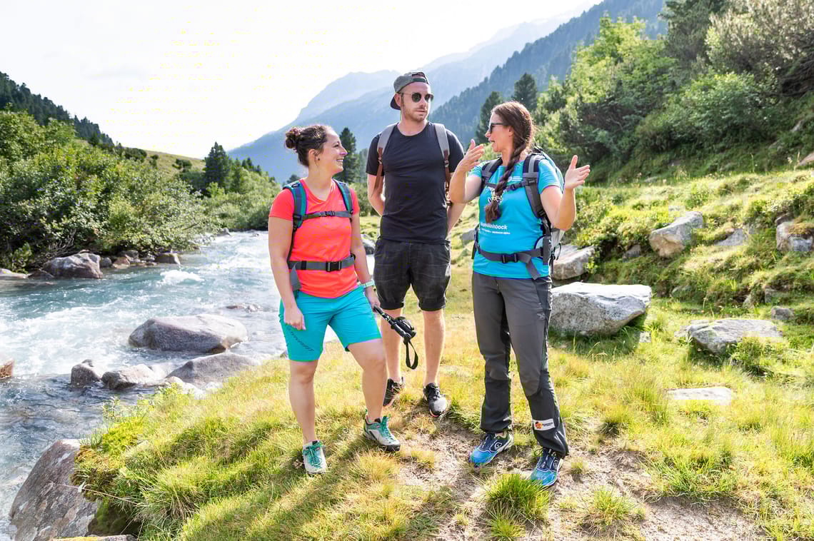 Guided Hike on the Schlegeis Three hikers with rucksacks walk along a path in the Zillertal Alps Nature Park in Schlegeis. Surrounded by green trees and a river, they enjoy the sunny landscape.