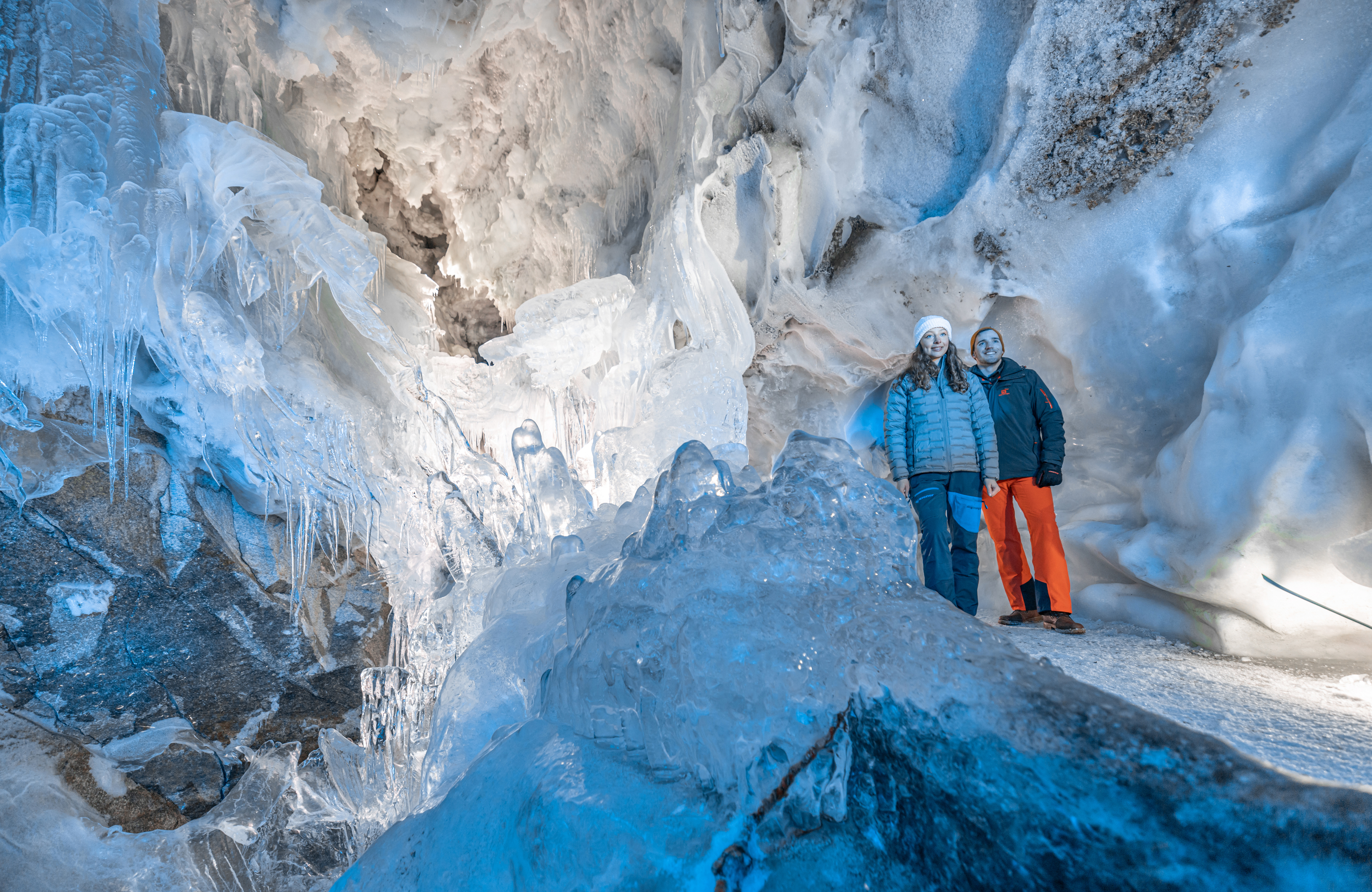 Zwei Besucher erkunden den Natur Eis Palast am Hintertuxer Gletscher: eine Person in blauer Jacke, eine in oranger Hose, umgeben von leuchtend blauen Eiskristallwänden, im Vordergrund ein Metallgeländer.