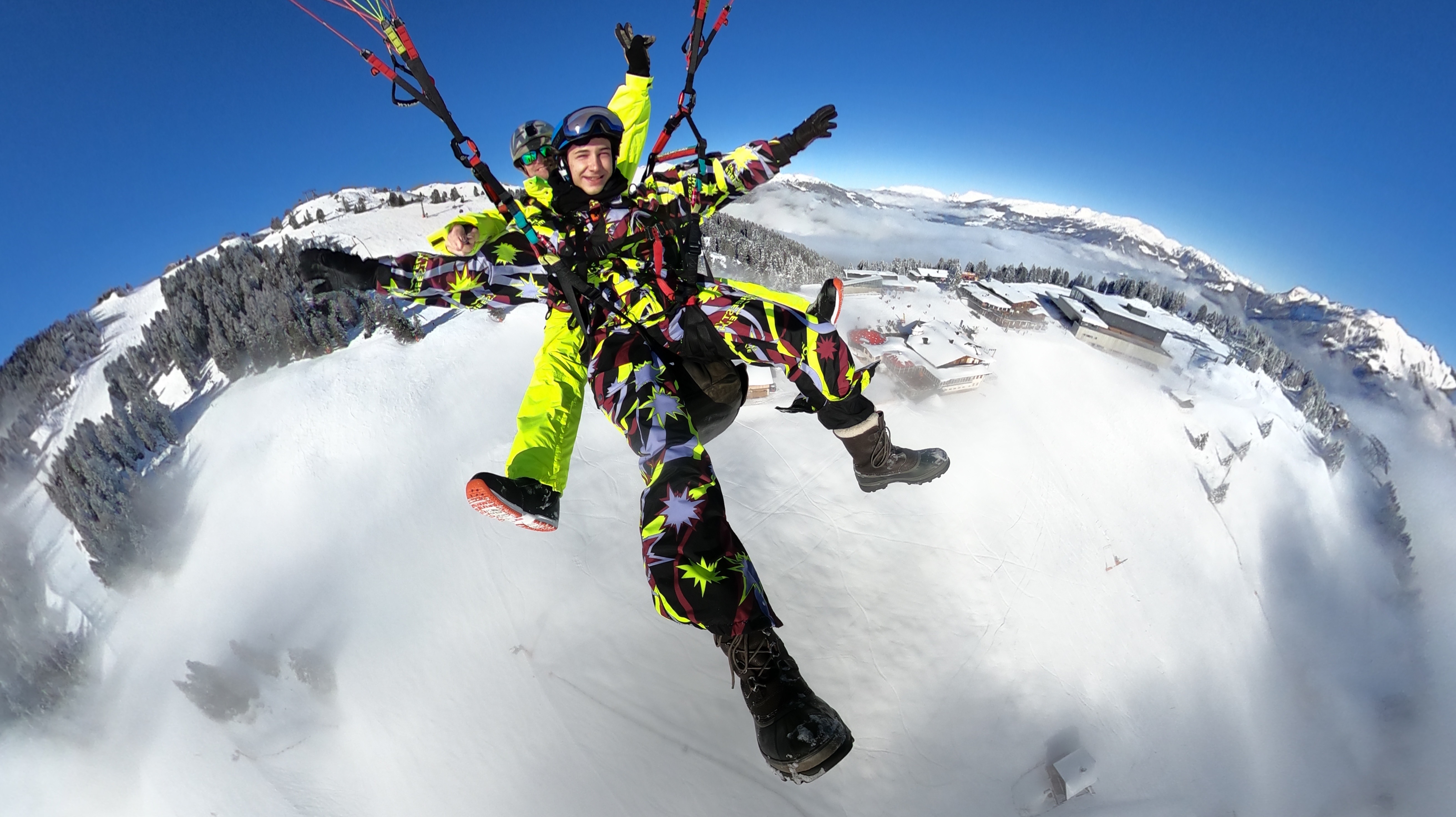 The picture shows two people paragliding high above the clouds. Both are wearing weatherproof clothing and helmets, enjoying the breathtaking view together. The sky is bright blue, and white clouds pass beneath them.