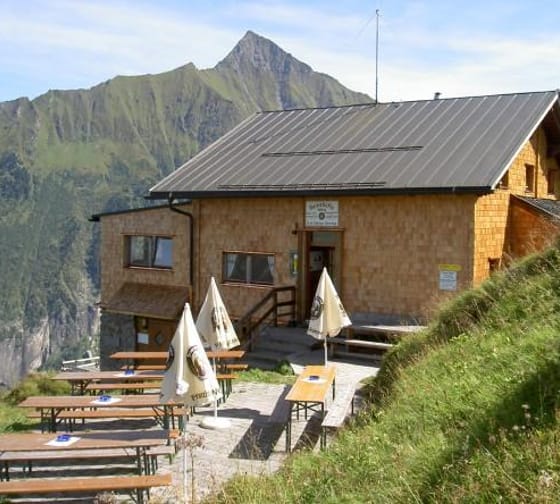 A cabin with outdoor tables and umbrellas, surrounded by green hills and mountains in the background.