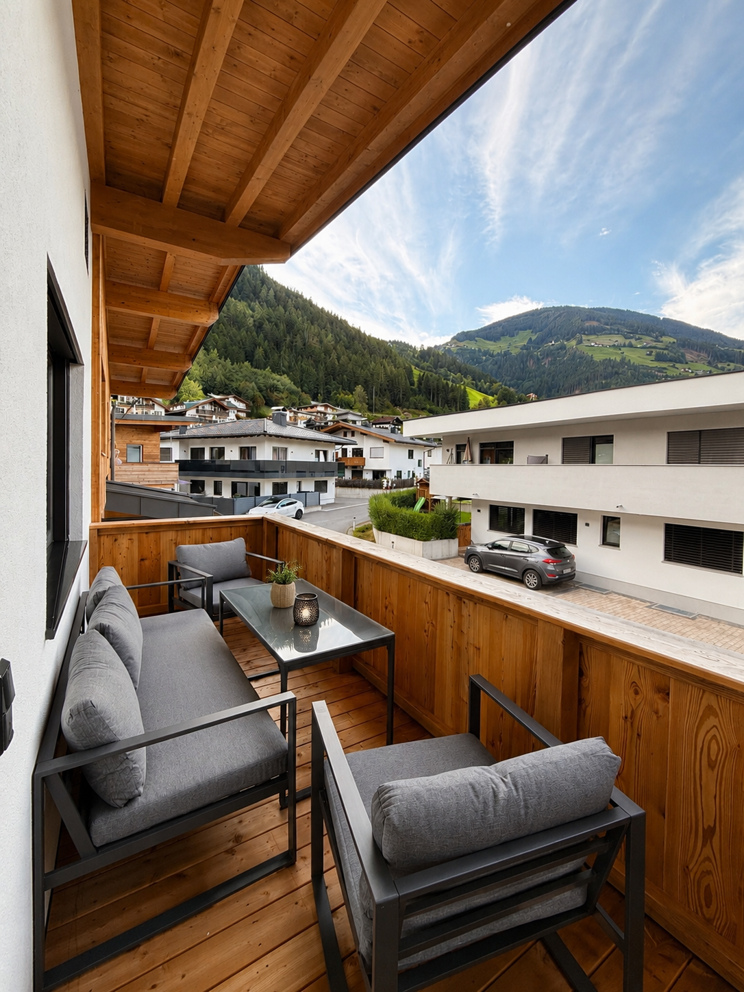 Balkon mit grauen Sitzmöbeln, Holzverkleidung, Blick auf Häuser und Berge bei schönem Himmel.