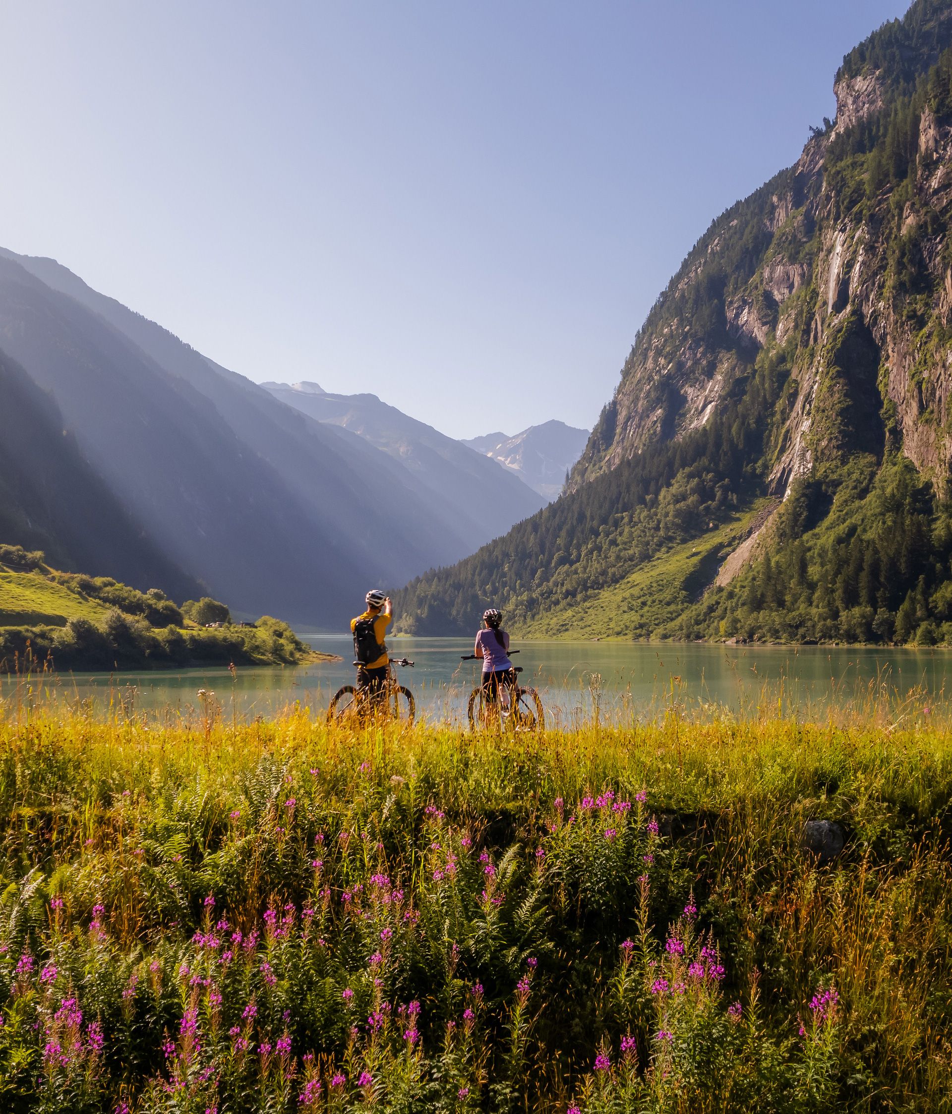 Sonnenaufgang im Naturparadies Stilluptal bei Mayrhofen im Zillertal: grüne Almwiese mit Blumen, zwei Wanderer auf Pfad, dahinter hohe Alpenberge und Felswände im weiten Tal.