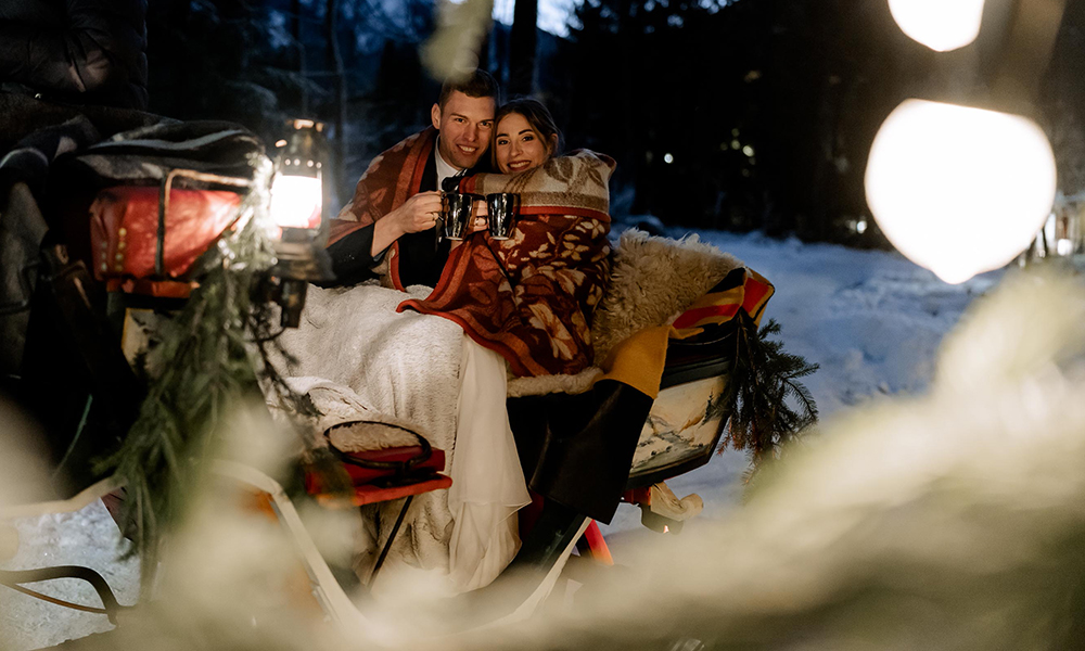 Ein Paar sitzt warm eingehüllt in Decken auf einer Pferdekutsche im Schnee. Laternenlicht leuchtet sanft, umgeben von winterlicher Landschaft und gemütlicher Stimmung. Romantische Winterfahrt durch die Nacht.