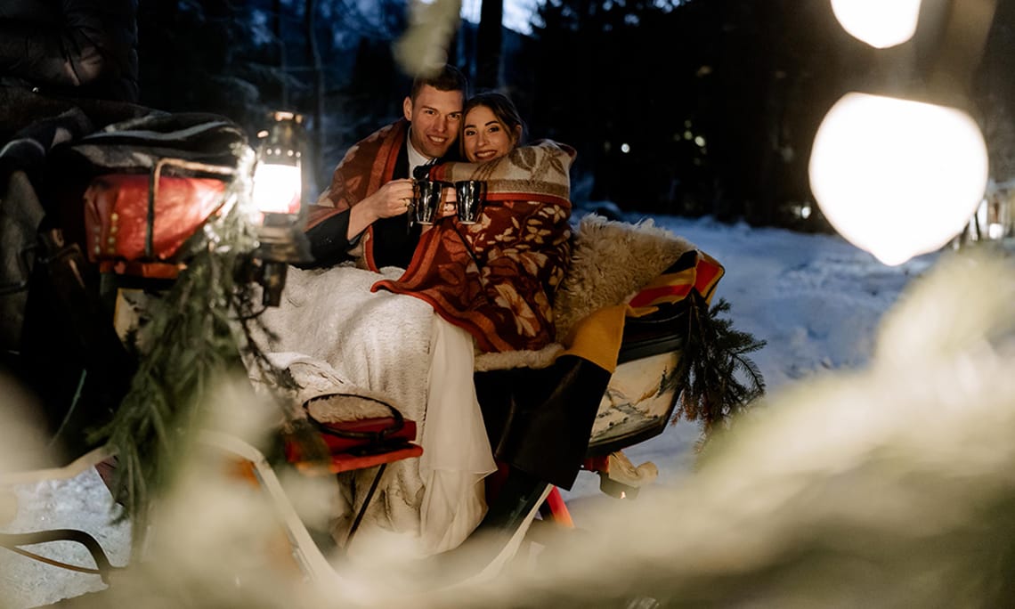 1. Adventtürchen - Zweisamkeit im Winterzauber Ein Paar sitzt warm eingehüllt in Decken auf einer Pferdekutsche im Schnee. Laternenlicht leuchtet sanft, umgeben von winterlicher Landschaft und gemütlicher Stimmung. Romantische Winterfahrt durch die Nacht.