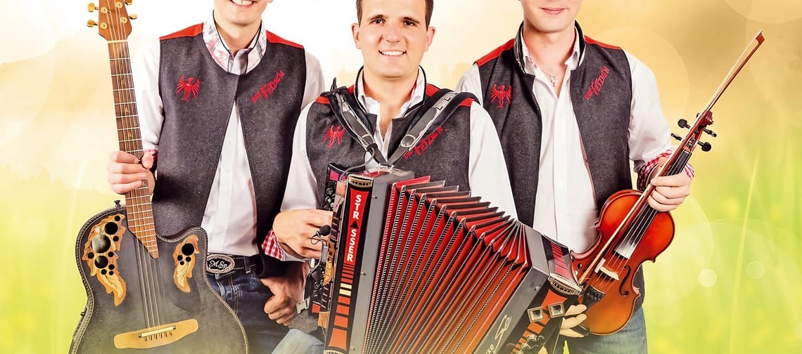 Drei Männer in Trachten mit Musikinstrumenten, lachend vor gelbem Hintergrund, feiern gemeinsam.