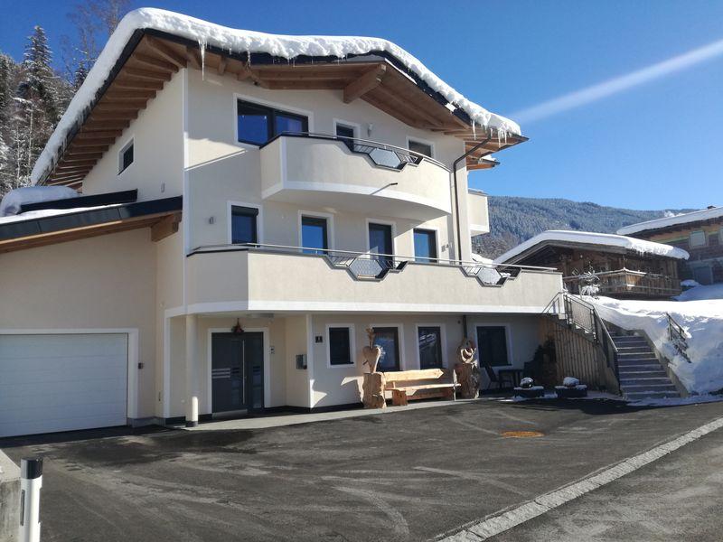 Modernes mehrstöckiges Haus mit Balkon, Schneebedecktem Dach und Holzskulpturen vor Eingang, bei sonnigem Himmel.