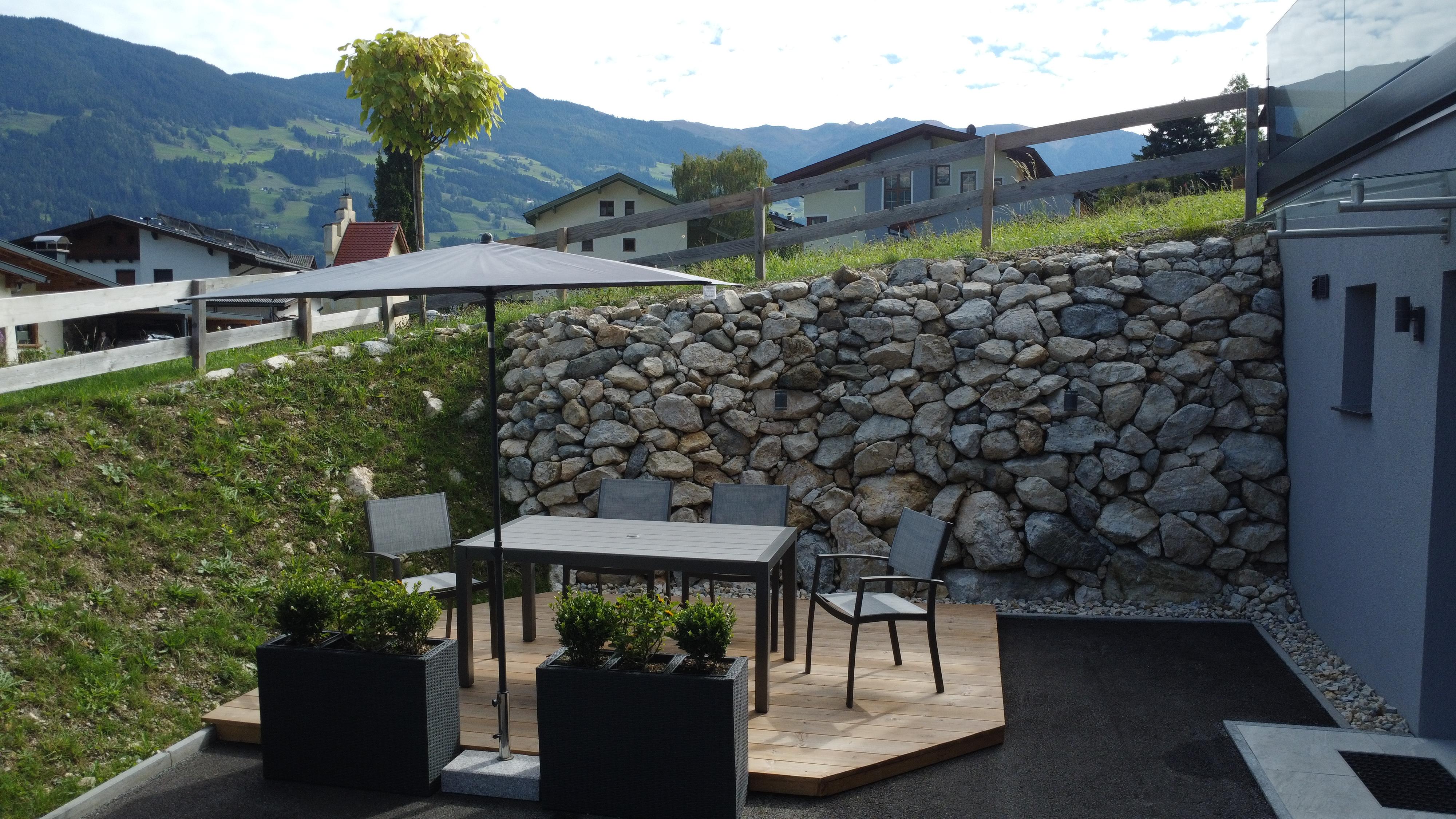 Garden with table, chairs, umbrella, stone wall, and mountain view in the background.