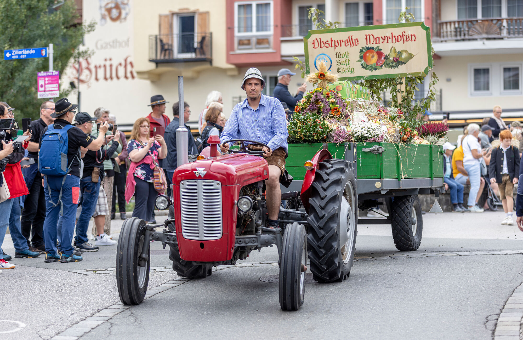 Brauchtumsumzug beim Zillertaler Bauern- und Musikherbst: Teilnehmer in traditioneller Tracht fahren mit einem roten Oldtimer-Traktor durch den Ort, während zahlreiche Zuschauer den Umzug verfolgen.