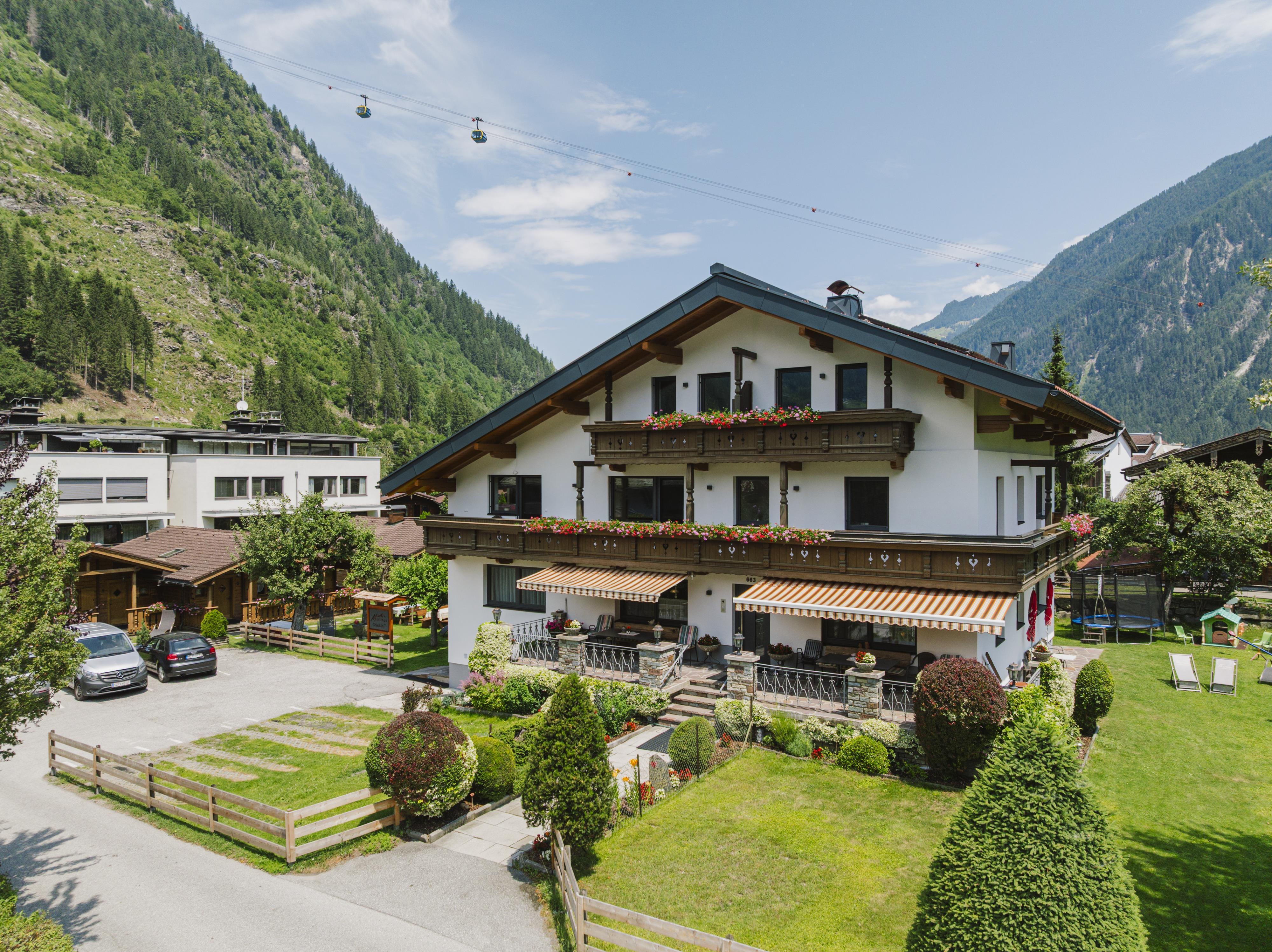 Ein Haus mit Balkon und Blumen vor, umgeben von grünen Gärten und Bergen im Hintergrund, bei schönem Wetter.