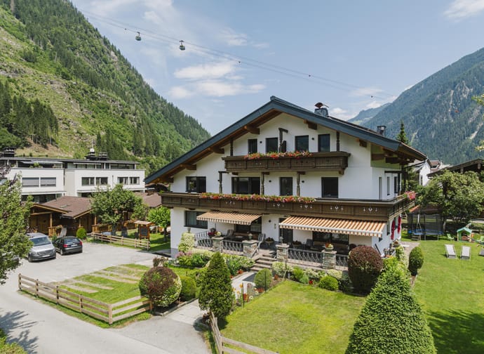 Ein Haus mit Balkon und Blumen vor, umgeben von grünen Gärten und Bergen im Hintergrund, bei schönem Wetter.