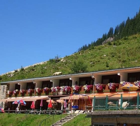 Mehrstöckiges Hotel mit Balkon, blumengeschmückten Töpfen und Sonnenschirmen vor einer grünen Berghügelkulisse.
