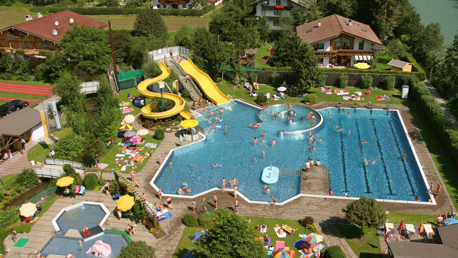 Aerial view of the outdoor swimming pool at Sommerwelt Hippach in the Zillertal valley with water slide, children's area, sports pool, and sunbathing lawn – a popular destination for families and swimming enthusiasts in Tyrol.