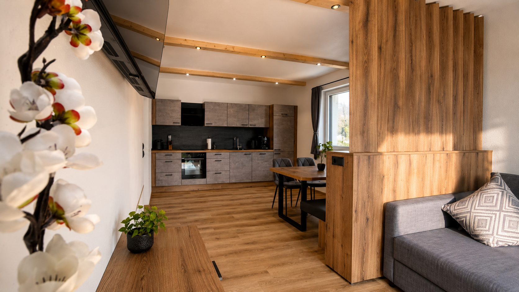 Moderner Wohnraum mit offener Küche, Holzdekor, grauem Sofa und Pflanzen, helle Fenster, warme Beleuchtung.