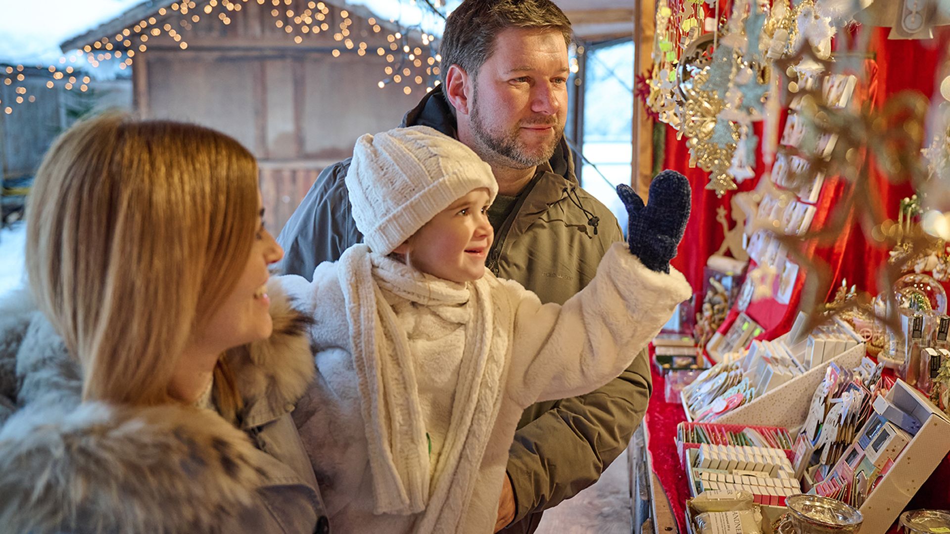 A family with a child visits a festively decorated stall at the Mayrhofner Advent. The child, dressed in white winter clothes, points enthusiastically at the colorful decorations while the parents smile.