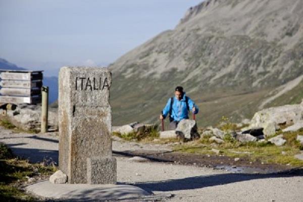 Hiker with backpack near border stone labeled 'ITALIA' in mountainous landscape.