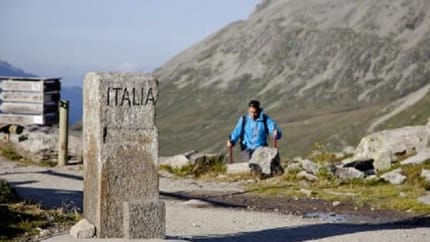 Hiker with backpack near border stone labeled 'ITALIA' in mountainous landscape.